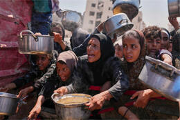 Palestinian women in headscarves with desperate looks on their faces push forward large pots at a food distribution centre in Gaza